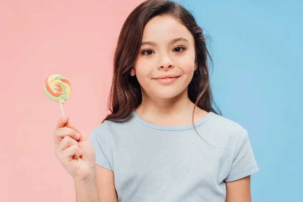 Entzückendes Kind, das Süßigkeiten hält, während es in die Kamera auf blauem und rosa Hintergrund schaut — Stockfoto