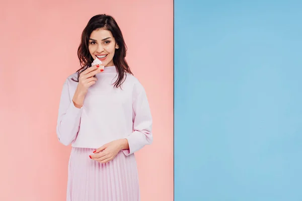 Schöne Frau isst Cupcake und schaut in die Kamera auf blauem und rosa Hintergrund — Stockfoto