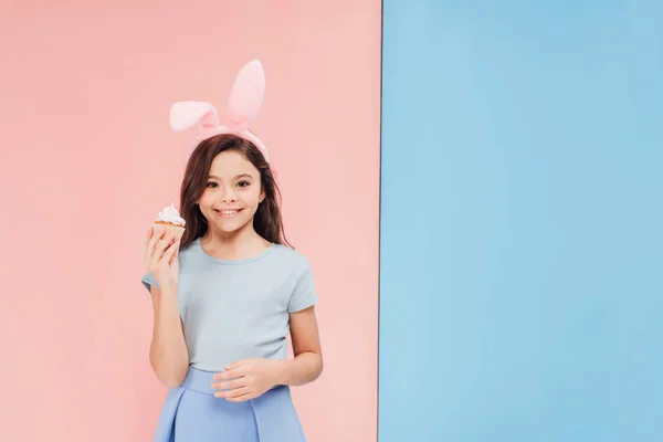 Adorabile bambino in orecchie di coniglio pasquale tenendo cupcake e guardando la fotocamera su sfondo blu e rosa — Foto stock