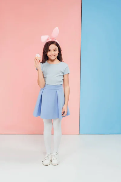 Adorable enfant dans les oreilles de lapin de Pâques tenant cupcake sur fond bleu et rose — Photo de stock