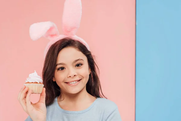 Adorable niño en conejito orejas celebración cupcake y mirando cámara en azul y rosa fondo - foto de stock