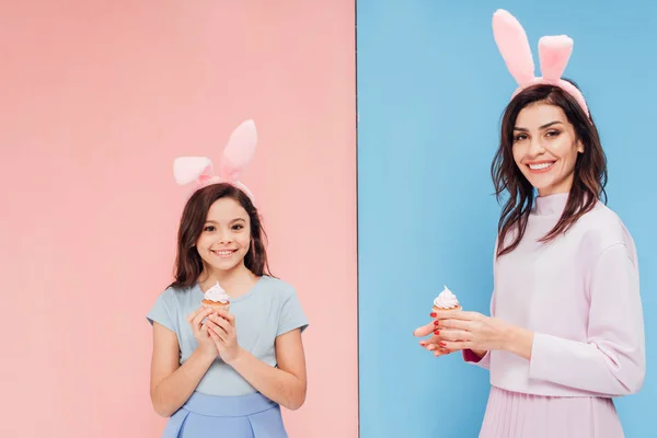 Hermosa mujer y niño en orejas de conejito sosteniendo cupcakes y mirando a la cámara sobre fondo azul y rosa - foto de stock