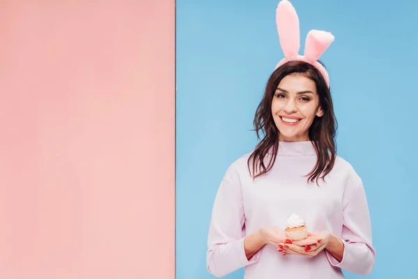 Bella donna in orecchie coniglietto pasquale tenendo cupcake e guardando la fotocamera su sfondo blu e rosa — Foto stock