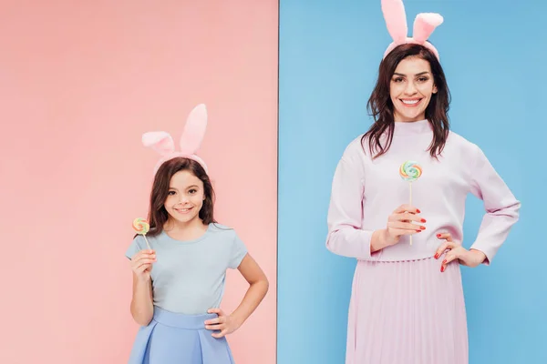 Schöne Frau und Kind in Hasenohren, die Bonbons halten, während sie in die Kamera auf blauem und rosa Hintergrund schauen — Stockfoto
