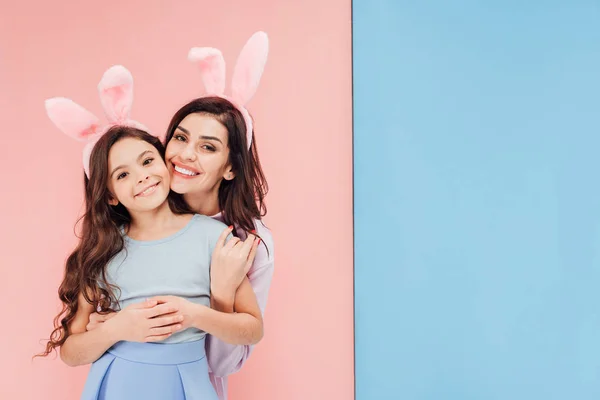 Schöne Frau in Osterhasenohren umarmt Kind und schaut in die Kamera auf blauem und rosa Hintergrund — Stockfoto