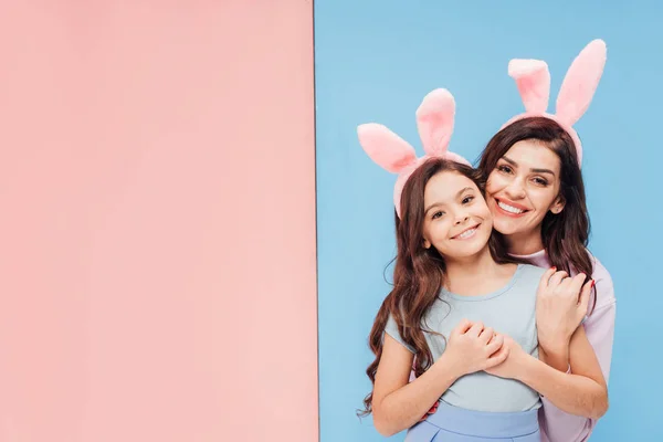 Hermosa mujer en orejas de conejo abrazando niño y sonriendo a la cámara sobre fondo azul y rosa - foto de stock