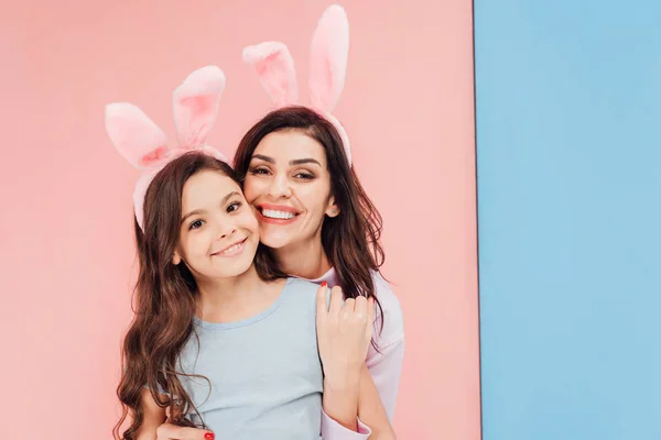 Bela mulher em easter bunny orelhas abraçando criança e olhando para a câmera no fundo azul e rosa — Fotografia de Stock