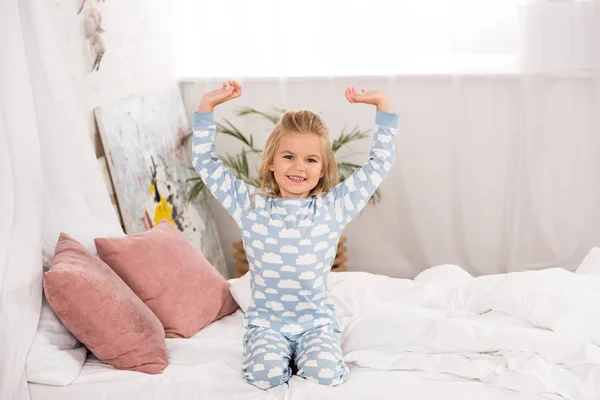 Улыбаясь милый ребенок в пижаме сидит на кровати с руками в воздухе — стоковое фото