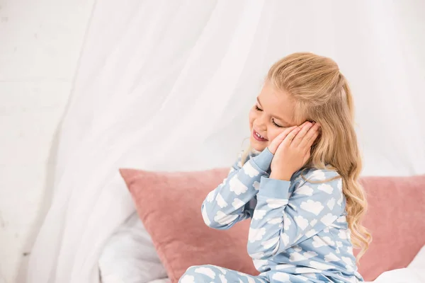 Lindo niño somnoliento sentado en la cama con los ojos cerrados y las manos cerca de las orejas - foto de stock