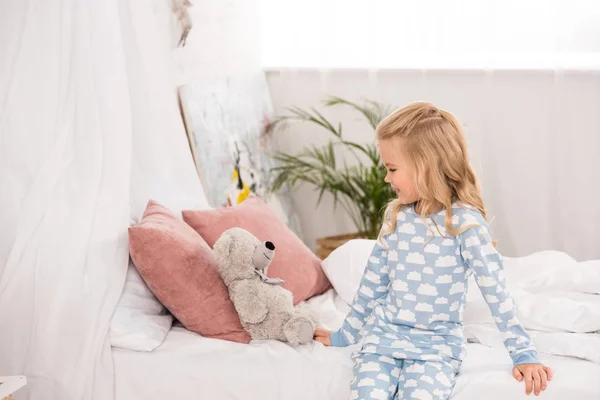 Cute adorable child in pajamas sitting on bed and looking at teddy bear — Stock Photo