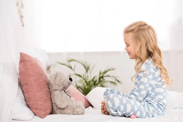 Vista lateral del lindo niño sentado en la cama con las piernas cruzadas y el oso de peluche - foto de stock