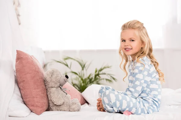 Niedliches Kind sitzt mit gekreuzten Beinen und Teddybär im Bett — Stockfoto