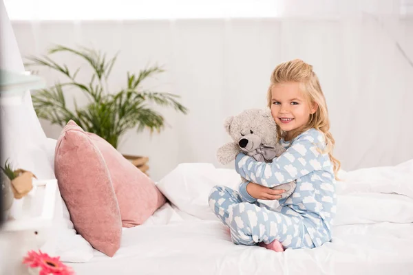 Niedliches Kind sitzt mit überkreuzten Beinen im Bett und umarmt Teddybär — Stockfoto