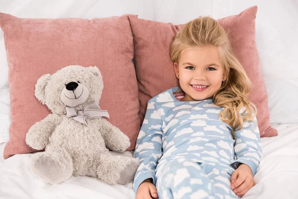 Liebevoll lächelndes Kind liegt mit Teddybär im Bett — Stockfoto