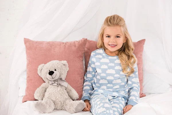 Мила усміхнена дитина сидить на ліжку з плюшевим ведмедем і подушками — стокове фото