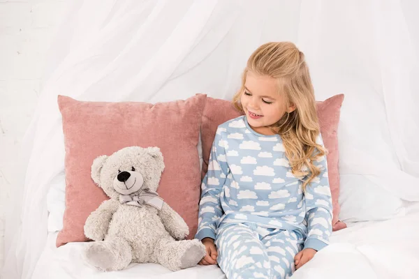 Bonito sorrindo criança sentado na cama e olhando para ursinho de pelúcia — Fotografia de Stock
