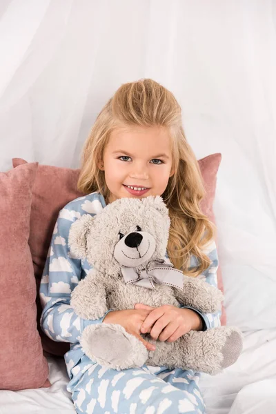 Garoto bonito alegre abraçando ursinho de pelúcia na cama — Fotografia de Stock