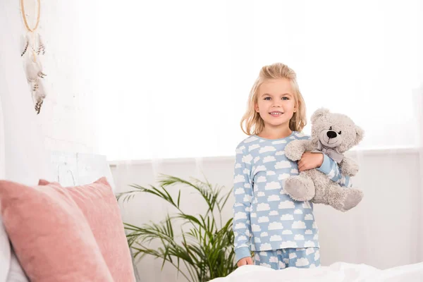 Sorrindo criança de pijama de pé com ursinho de pelúcia no quarto — Fotografia de Stock