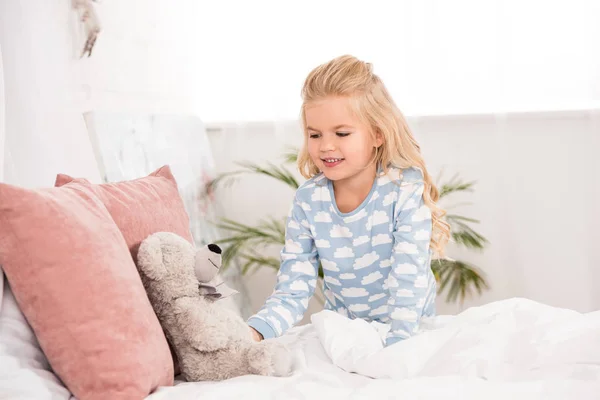 Cute child in pajamas looking at teddy bear in bed — Stock Photo