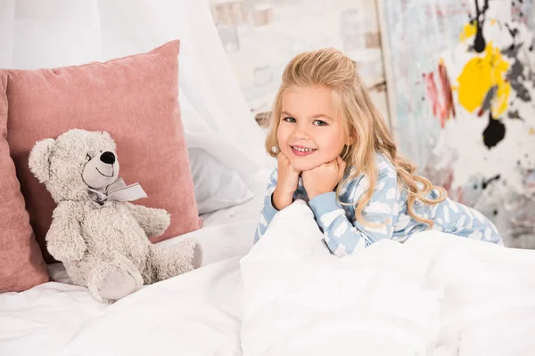 Criança adorável com ursinho na cama olhando para a câmera — Fotografia de Stock