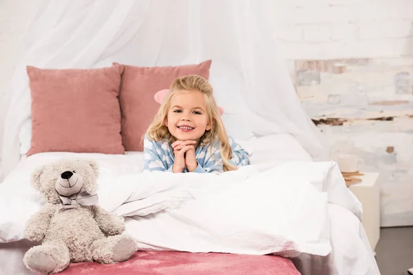 Entzückendes Kind liegt mit Teddybär im Bett und blickt in die Kamera — Stockfoto