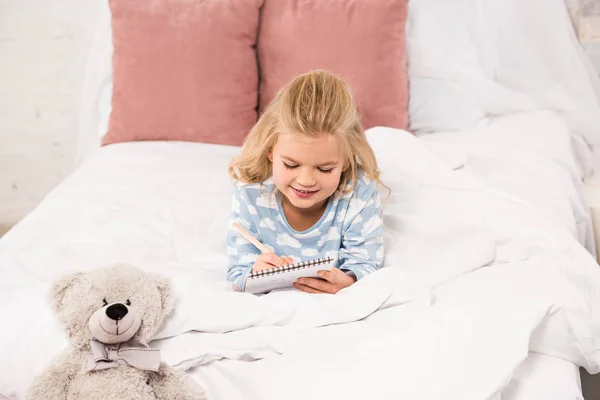 Adorabile bambino sdraiato a letto e scrivere nel taccuino — Foto stock