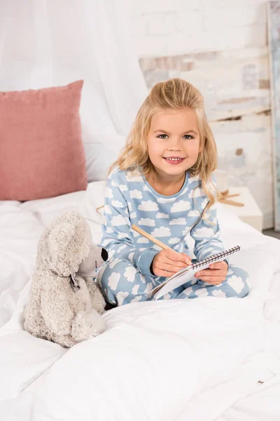 Adorable enfant assis au lit et écrivant dans un cahier — Photo de stock