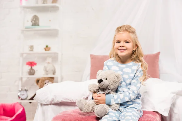 Lächelndes süßes Kind sitzt mit Teddybär im Bett und schaut in die Kamera — Stockfoto