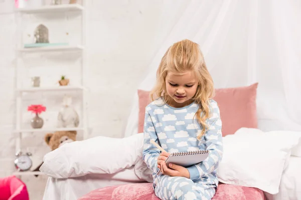 Nettes Kind im Schlafanzug sitzt auf dem Bett und schreibt in Notizbuch — Stockfoto