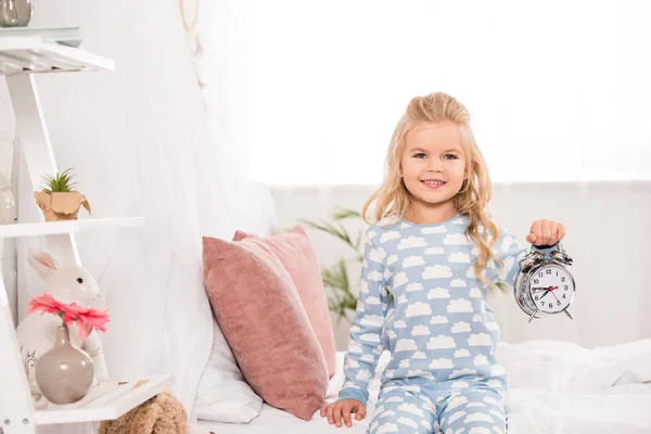 Lächelndes liebenswertes Kind, das mit Uhr auf dem Bett sitzt — Stockfoto