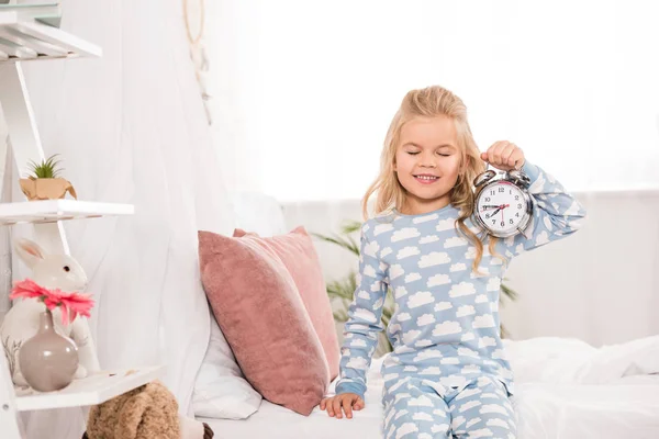 Happy adorable kid sitting on bed with clock and closed eyes — Stock Photo