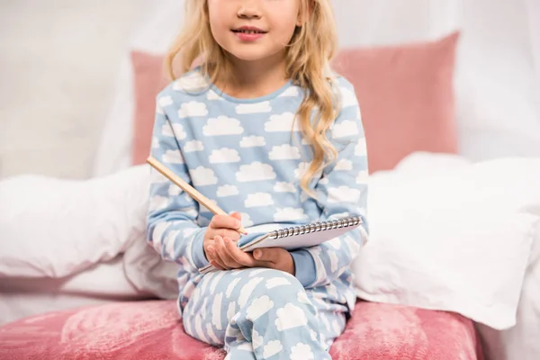 Обрізаний вид дитини в піжамі сидить на ліжку і пише в блокноті — стокове фото