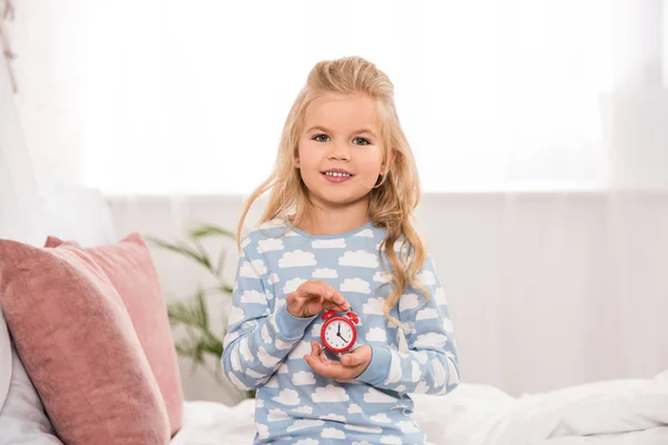 Glückliches liebenswertes Kind sitzt auf dem Bett mit kleiner Uhr in den Händen — Stockfoto