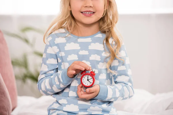 Vue partielle de l'adorable enfant assis sur le lit avec petite horloge dans les mains — Photo de stock