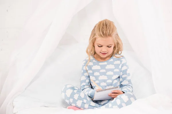 Чарівна дитина в піжамі сидить на ліжку з схрещеними ногами і пише в блокноті — стокове фото