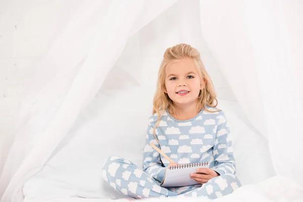 Entzückendes Kind im Schlafanzug, das in Notizbuch schreibt und in die Kamera schaut — Stockfoto