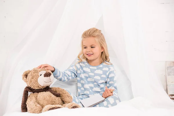 Garoto bonito em pijama sentado na cama com notebook e ursinho de pelúcia — Fotografia de Stock