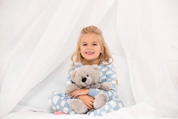 Adorable child in pajamas sitting on bed and embracing teddy bear — Stock Photo