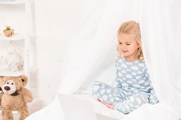 Niño sonriente sentado en la cama en pijama y usando el ordenador portátil - foto de stock