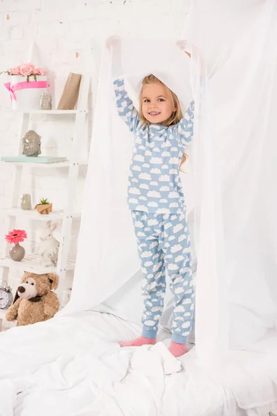 Улыбающийся ребенок в пижаме стоит на кровати и трогает навес — стоковое фото