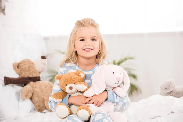 Niño adorable sonriente sentado con juguetes suaves en la cama - foto de stock