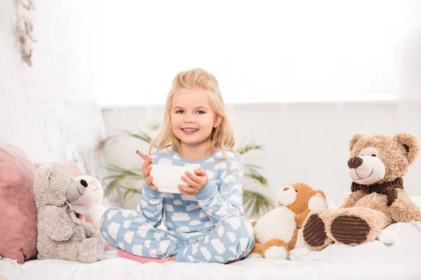 Enfant souriant assis au lit avec des jouets mous et tenant bol avec petit déjeuner — Photo de stock