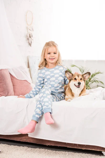 Criança sorridente feliz em pijama sentado com pernas cruzadas e cão corgi na cama — Fotografia de Stock