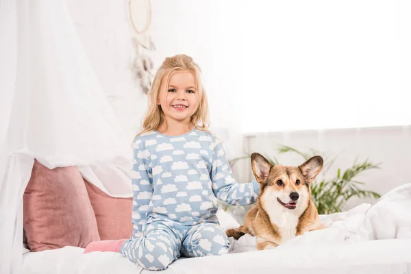 Счастливый улыбающийся ребенок в пижаме, сидящий с собакой корги в постели — стоковое фото