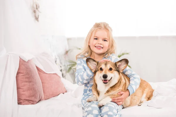 Bambino sorridente in pigiama che abbraccia e accarezza il cane corgi a letto — Foto stock