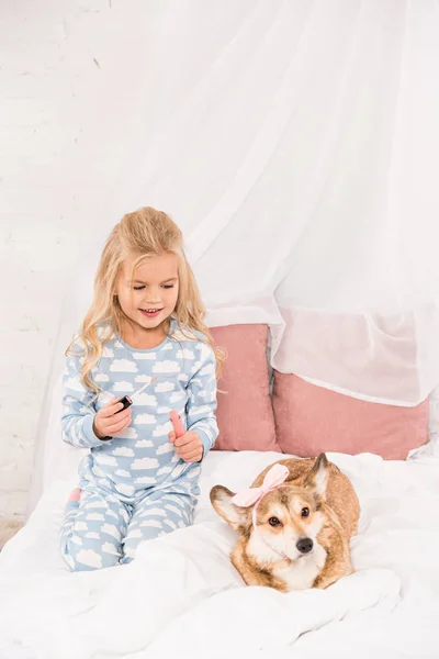 Bonito criança sentado na cama com corgi cão e segurando batom gloss — Fotografia de Stock