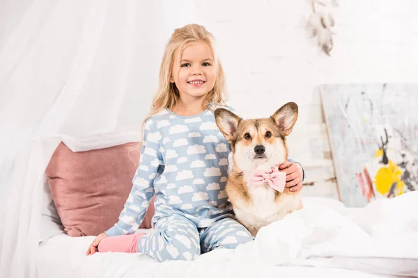 Niño sonriente sentado en la cama con pembroke perro corgi galés con arco en casa - foto de stock