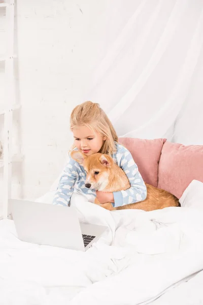 Adorabile bambino sdraiato a letto, abbracciando cane corgi e utilizzando il computer portatile a casa — Foto stock