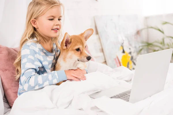 Sorrindo criança deitada na cama, abraçando cão corgi e usando laptop em casa — Fotografia de Stock