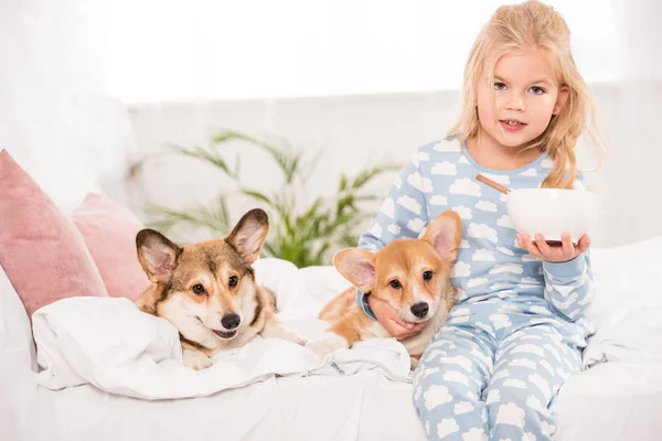 Mignon enfant assis sur le lit avec des chiens gallois corgi pembroke et tenant bol de céréales — Photo de stock